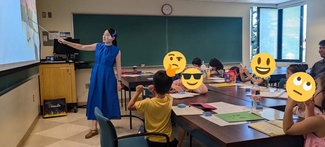 A Chinese teacher in a blue dress gestures at a screen at the front of a classroom as several young students look on.