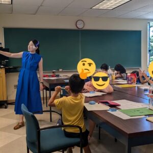 A Chinese teacher in a blue dress gestures at a screen at the front of a classroom as several young students look on.