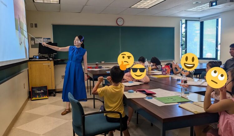 A Chinese teacher in a blue dress gestures at a screen at the front of a classroom as several young students look on.